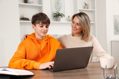 Smiling mother and her son doing homework with laptop at table indoors