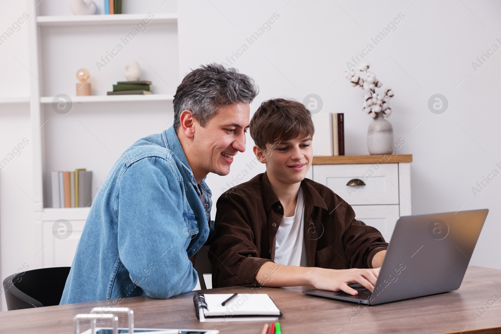 Photo of Smiling father and his son doing homework with laptop at table indoors