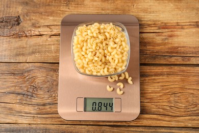 Photo of Modern electronic kitchen scale with bowl of raw pasta on wooden table, top view