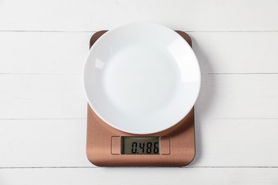 Photo of Modern electronic kitchen scale with plate on white wooden table, top view