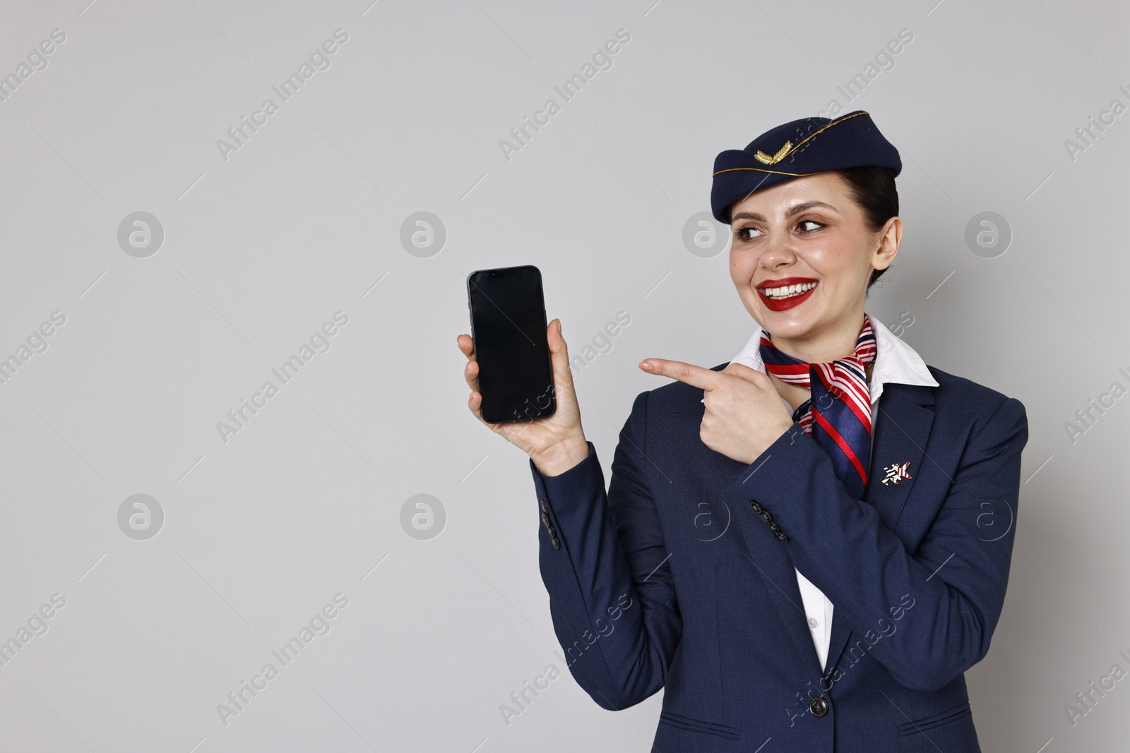 Photo of Smiling stewardess pointing at smartphone on grey background. Space for text