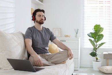 Photo of Handsome man in headphones meditating near laptop on sofa at home