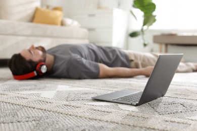 Photo of Man in headphones meditating on floor at home, focus on laptop