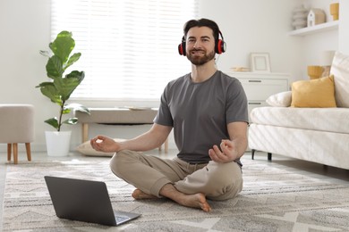 Photo of Smiling man in headphones meditating near laptop on floor at home