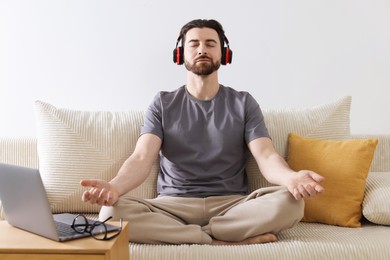 Photo of Man in headphones meditating near laptop on sofa at home