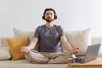 Photo of Man in headphones meditating near laptop on sofa at home