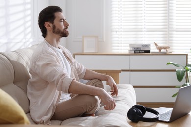 Photo of Man meditating near laptop on sofa at home