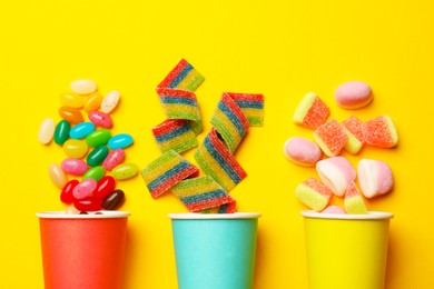 Photo of Tasty candies, marshmallows and paper cups on yellow background, flat lay