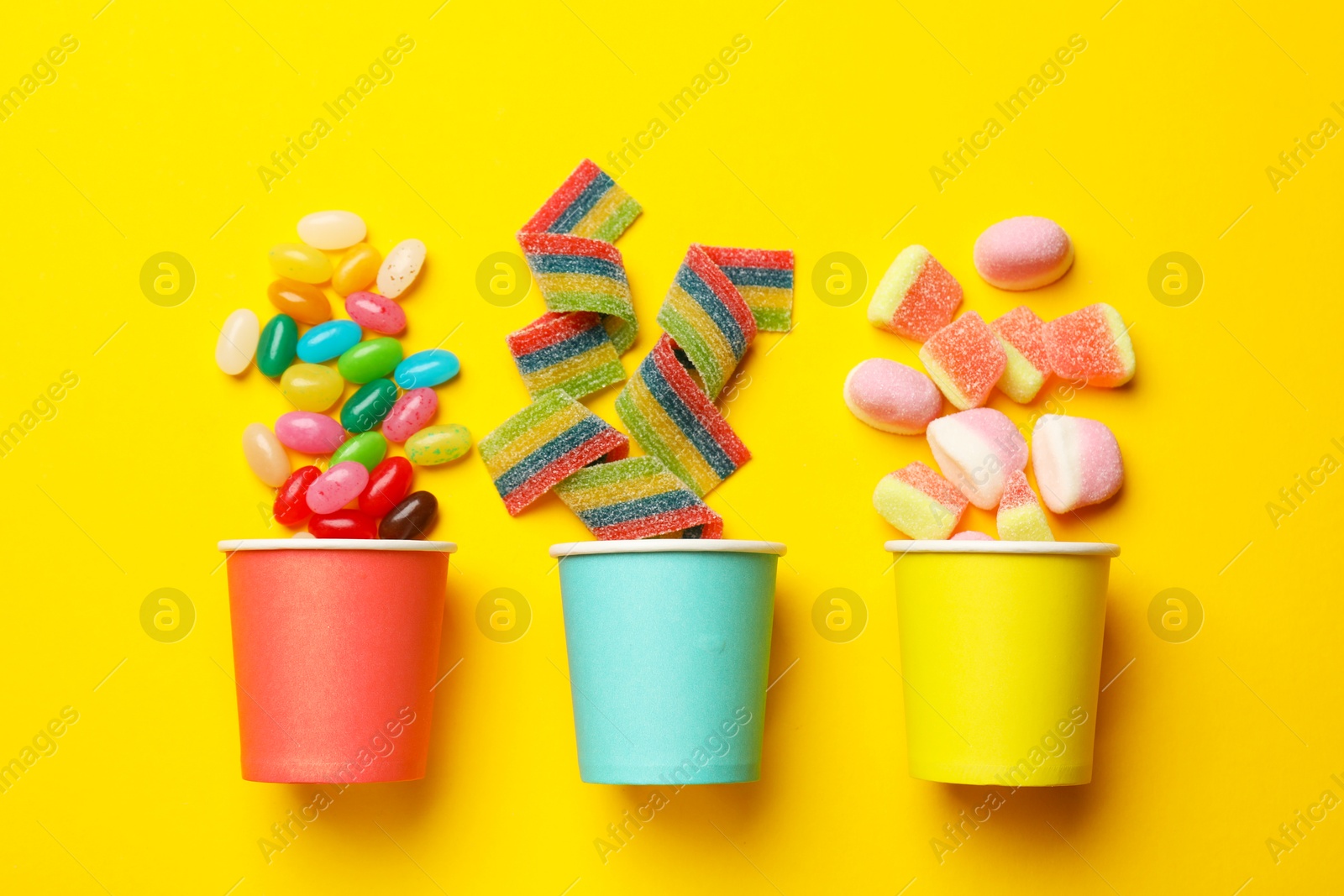 Photo of Tasty candies, marshmallows and paper cups on yellow background, flat lay