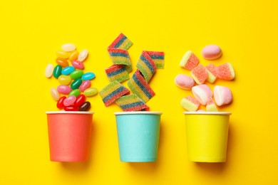 Tasty candies, marshmallows and paper cups on yellow background, flat lay