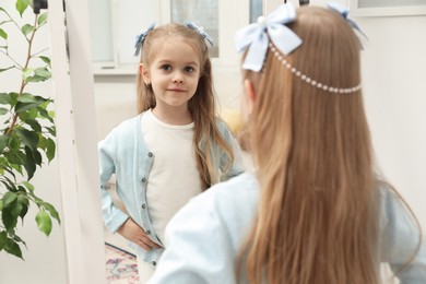 Photo of Cute little girl with beautiful hair accessory looking in mirror at home