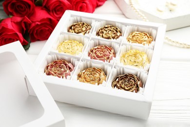 Photo of Flower shaped chocolate bonbons in box, red roses and pearl jewelry on white wooden table, closeup