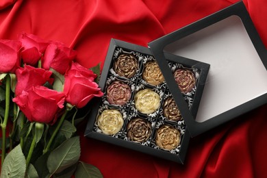 Photo of Delicious flower shaped chocolate bonbons in box and roses on red fabric background, top view