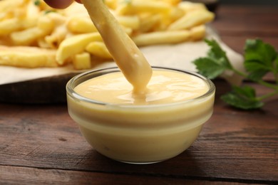 Photo of Dipping French fry into cheese sauce at wooden table, closeup