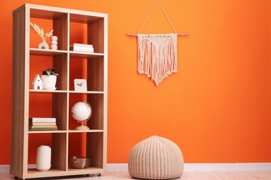 Photo of Wooden shelving unit with different home decor and pouf near orange wall indoors, space for text