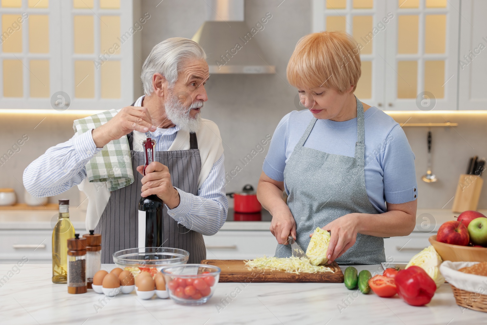 Photo of Senior man opening bottle of wine while cooking with his wife in kitchen