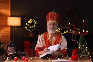 Saint Nicholas with notebook at desk in room decorated for Christmas