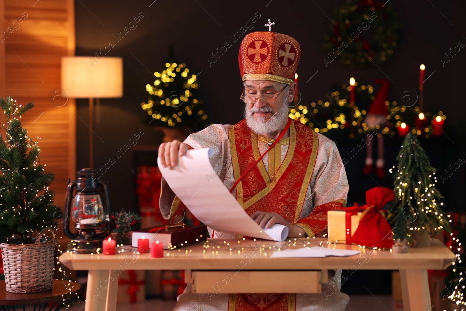 Photo of Saint Nicholas with gift list at desk in room decorated for Christmas