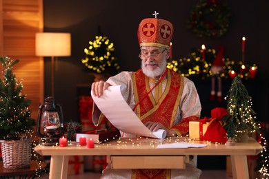 Saint Nicholas with gift list at desk in room decorated for Christmas