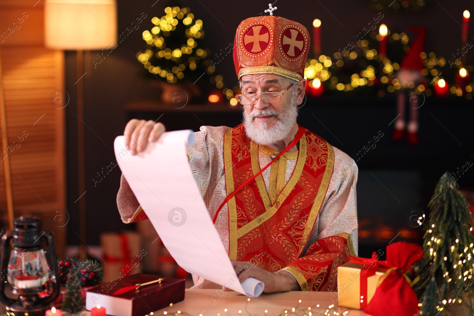 Photo of Saint Nicholas with gift list at desk in room decorated for Christmas