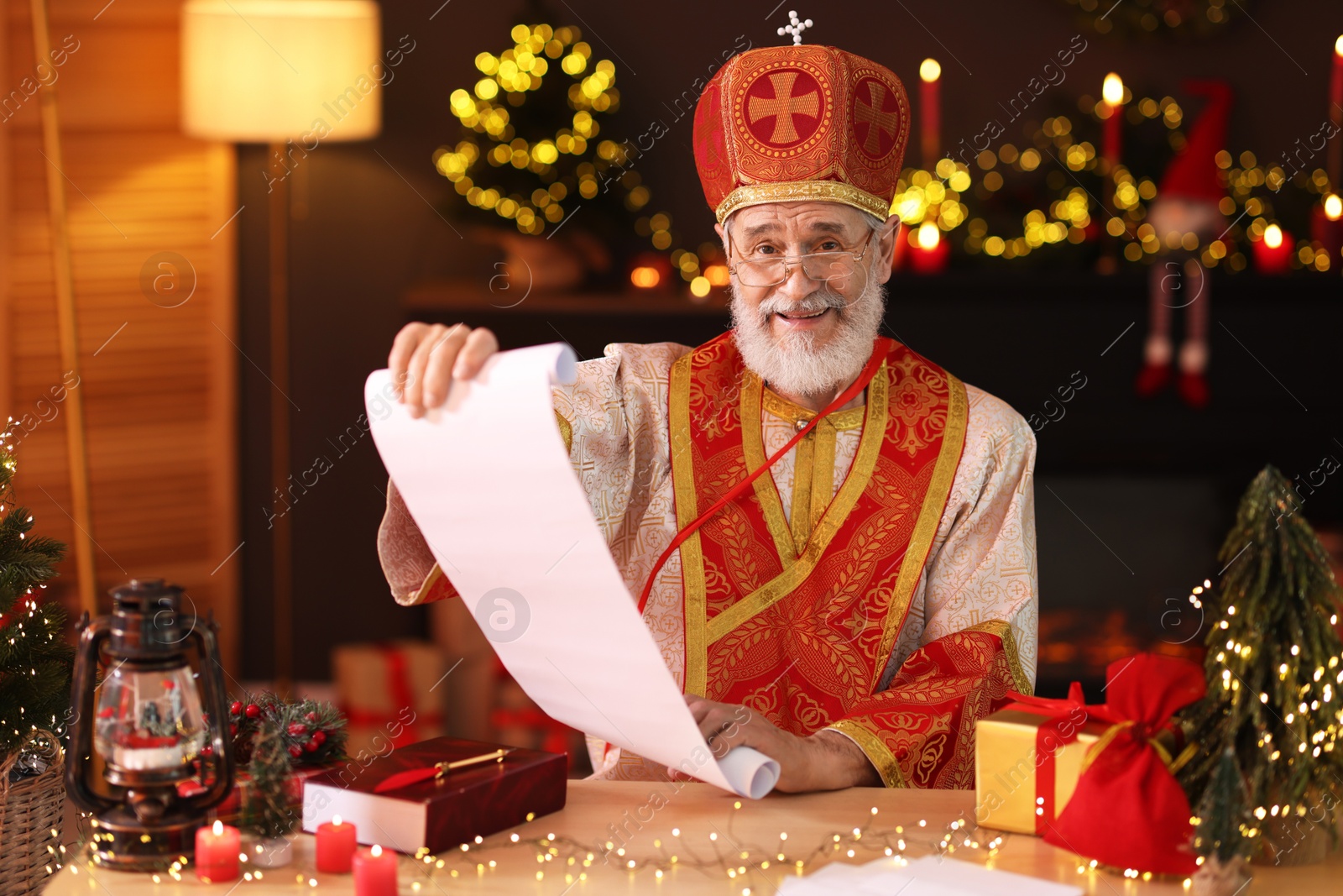 Photo of Saint Nicholas with gift list at desk in room decorated for Christmas