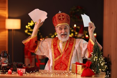 Saint Nicholas with letters at desk in room decorated for Christmas