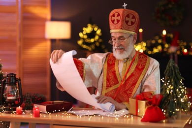 Photo of Saint Nicholas with gift list at desk in room decorated for Christmas