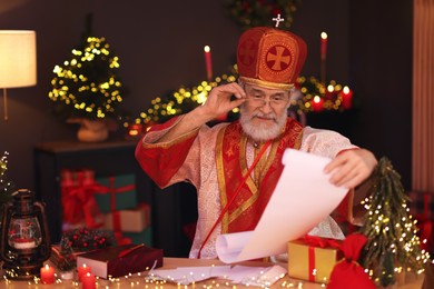 Saint Nicholas with gift list at desk in room decorated for Christmas