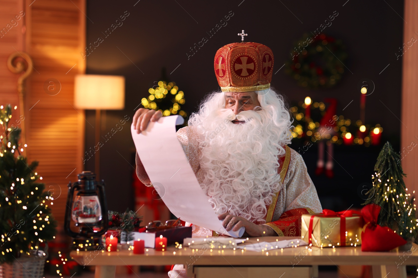 Photo of Saint Nicholas with gift list at desk in room decorated for Christmas