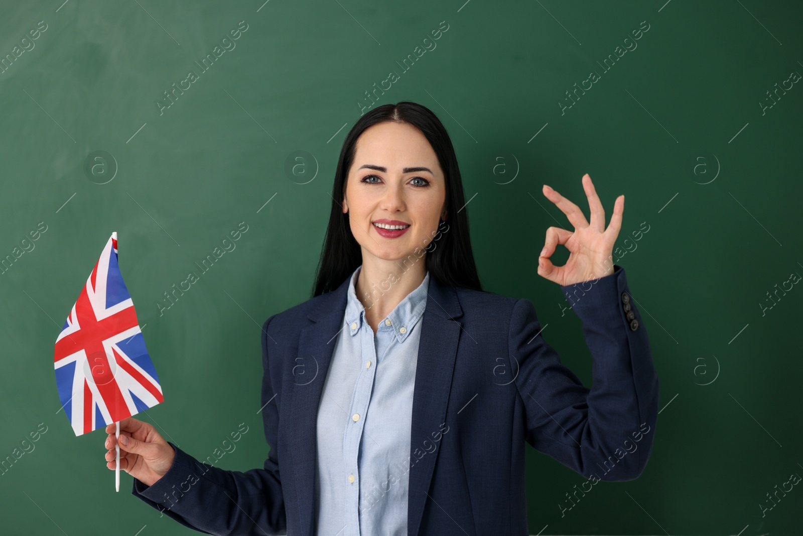 Photo of English teacher with UK flag showing ok gesture near chalkboard in classroom