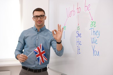 Photo of English teacher with UK flag showing OK gesture near whiteboard in classroom