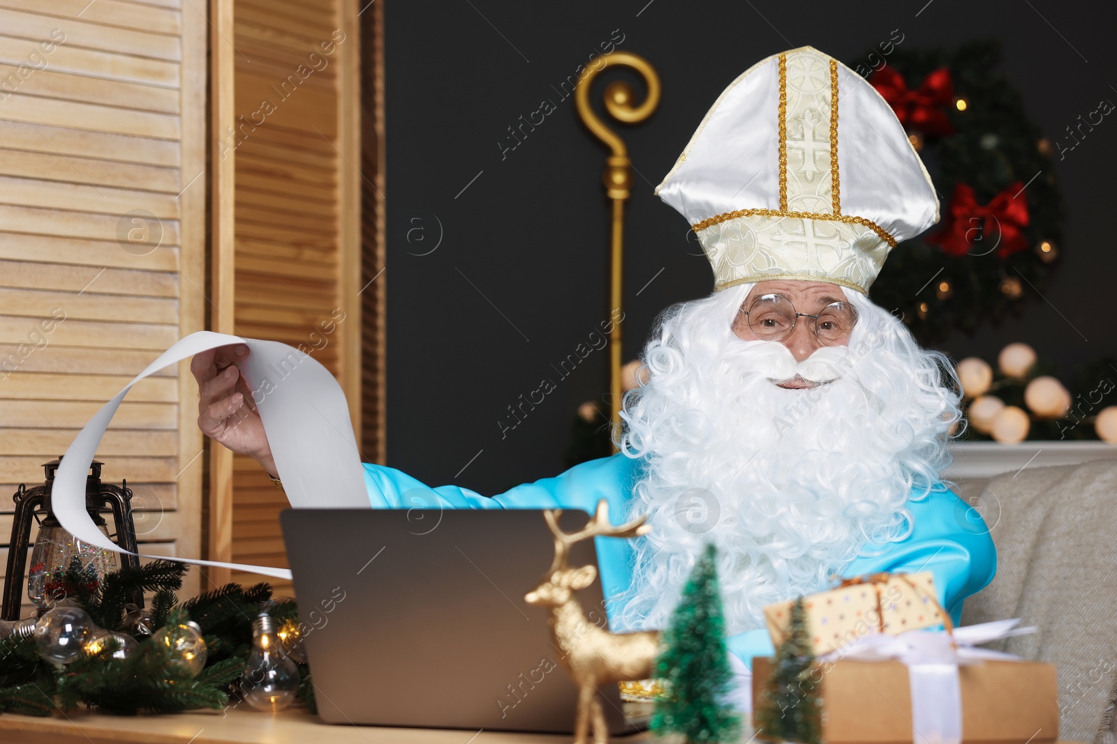 Photo of Saint Nicholas with wish list and laptop in room decorated for Christmas