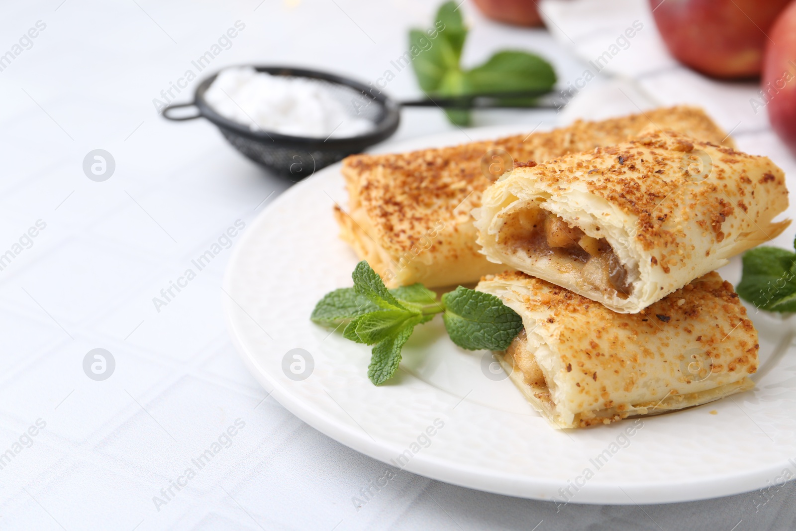 Photo of Tasty apple strudels mint and powdered sugar on white tiled table, closeup. Space for text