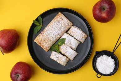 Photo of Tasty apple strudels with powdered sugar, mint and fruits on yellow background, flat lay