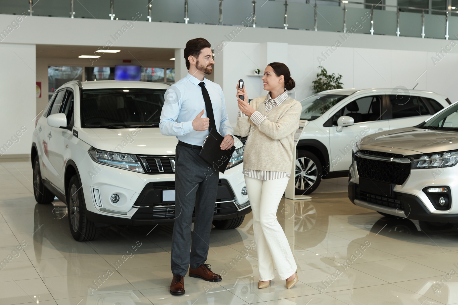 Photo of Happy salesman showing thumbs up and client with key near new car in salon