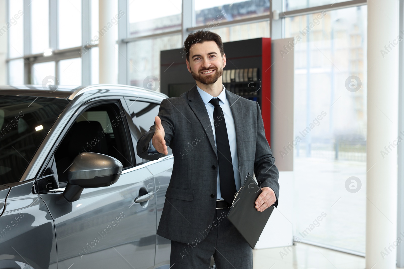 Photo of Happy salesman with clipboard offering handshake near new car in salon