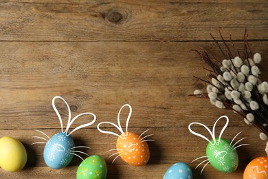 Image of Bright Easter eggs and willow branches on wooden table, top view. Some of them with bunny ears, noses and whiskers