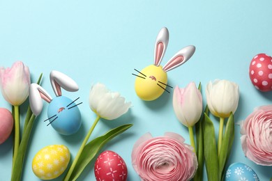 Image of Bright Easter eggs and flowers on light blue background, top view. Some of them with bunny ears, noses and whiskers