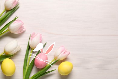 Image of Bright Easter eggs and flowers on white wooden table, top view. PInk one with bunny ears, nose and whiskers