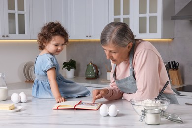 Photo of Cute little girl with her granny cooking by recipe book in kitchen