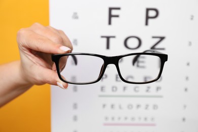 Photo of Woman holding glasses against vision test chart on orange background, closeup