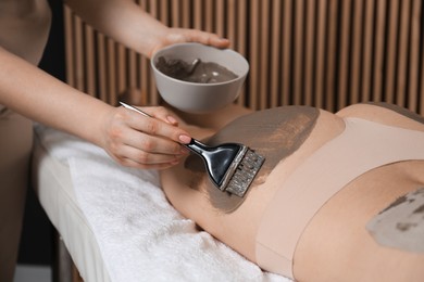 Photo of Esthetician applying cosmetic product for body wraps treatment onto woman's buttocks in spa salon, closeup