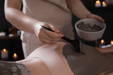 Photo of Esthetician applying cosmetic product for body wraps treatment onto woman's buttocks in spa salon, closeup