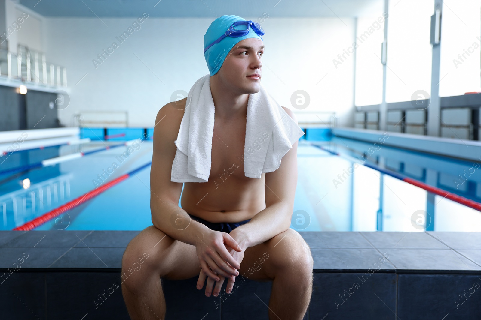 Photo of Young man wearing cap and goggles with towel near swimming pool indoors