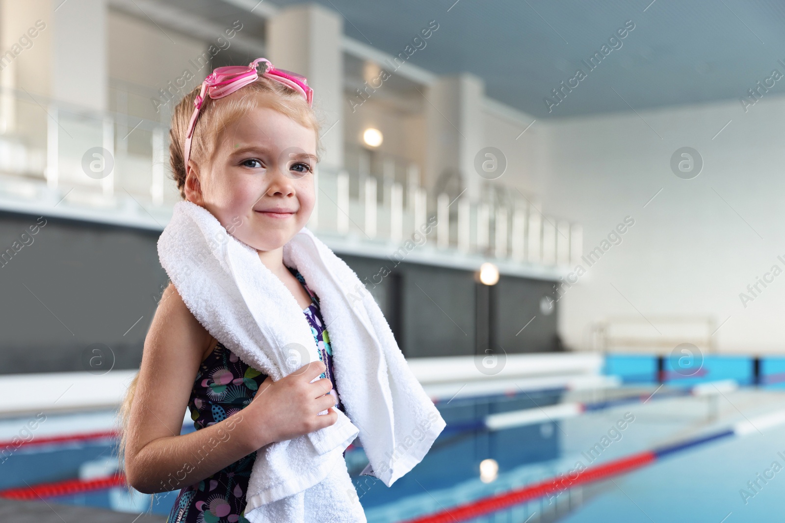 Photo of Little girl with goggles and towel near swimming pool indoors, space for text