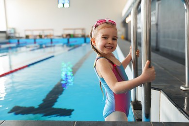 Little girl with goggles getting out of swimming pool indoors, space for text