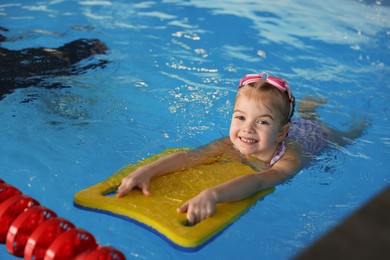 Photo of Little girl with goggles and kickboard swimming in pool indoors