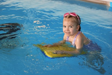 Photo of Little girl with goggles and kickboard swimming in pool indoors