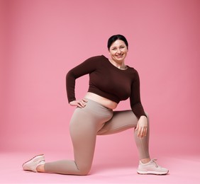 Plus size woman in gym clothes exercising on pink background