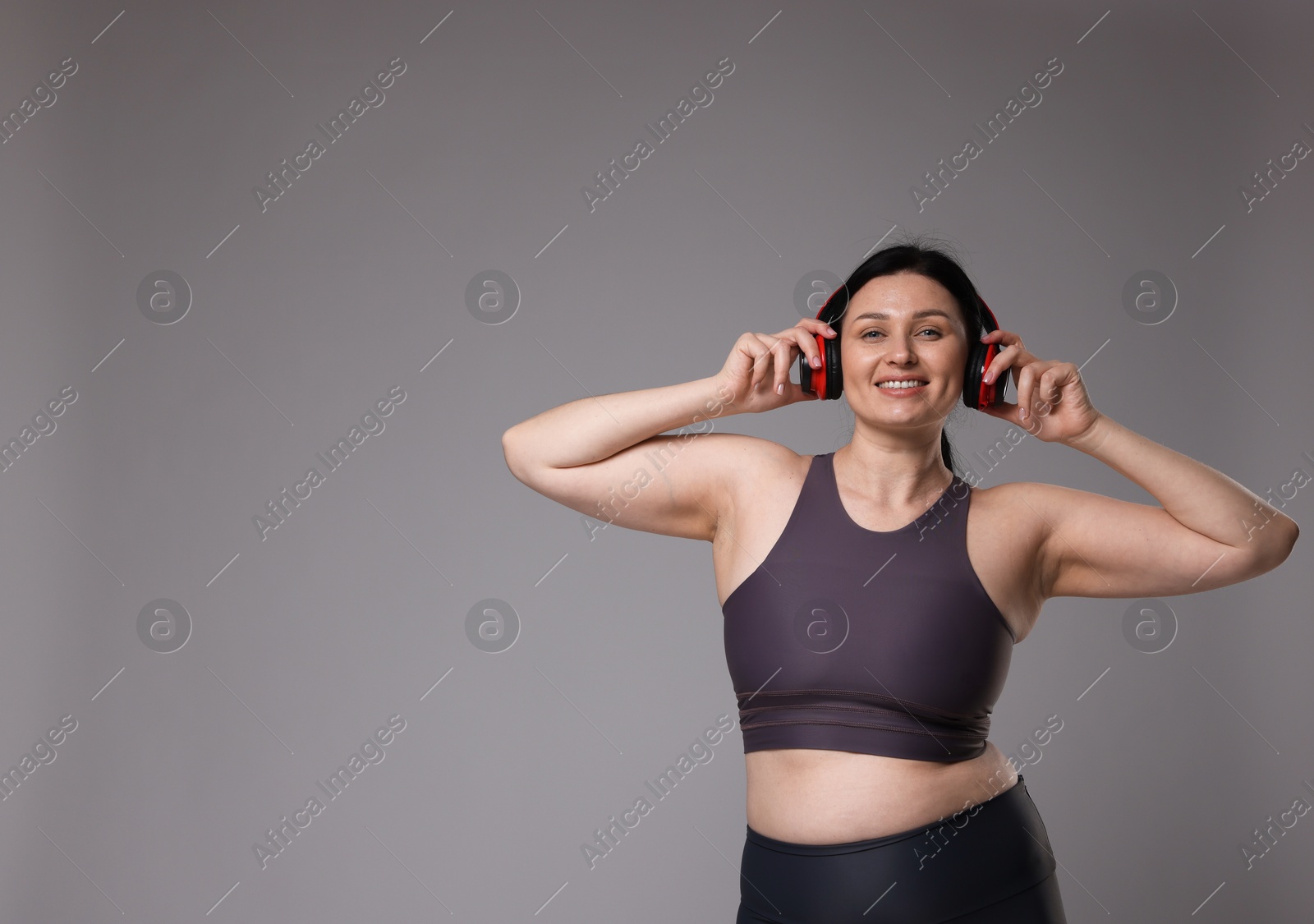 Photo of Plus size woman in gym clothes and headphones on grey background, space for text